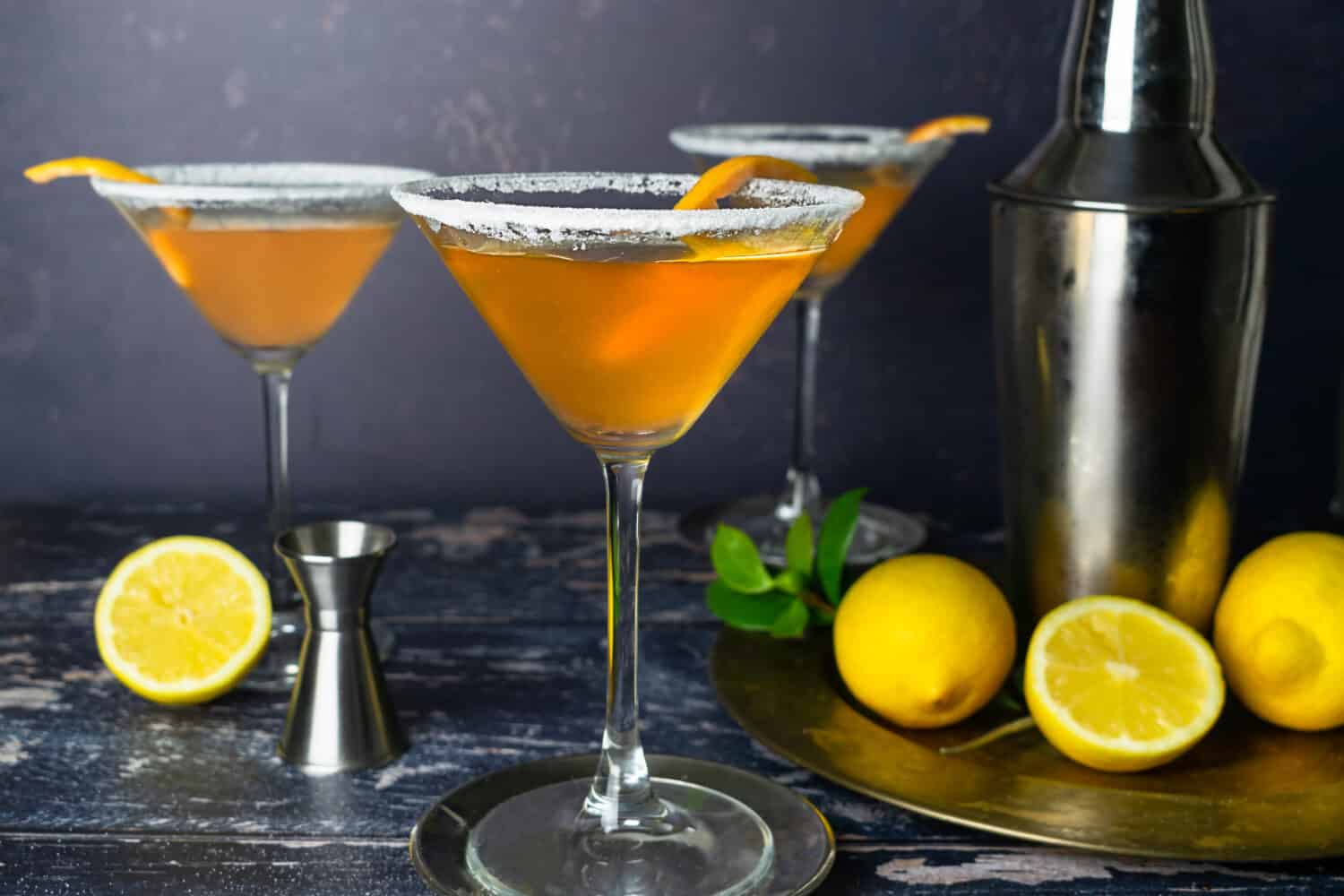Sidecar Cocktail in a Martini glass with an orange peel on a dark background. Ingredients: Cognac, Cointreau, lemon juice, sugar and ice. Shaker and lemon in the background. Sugar on the rim
