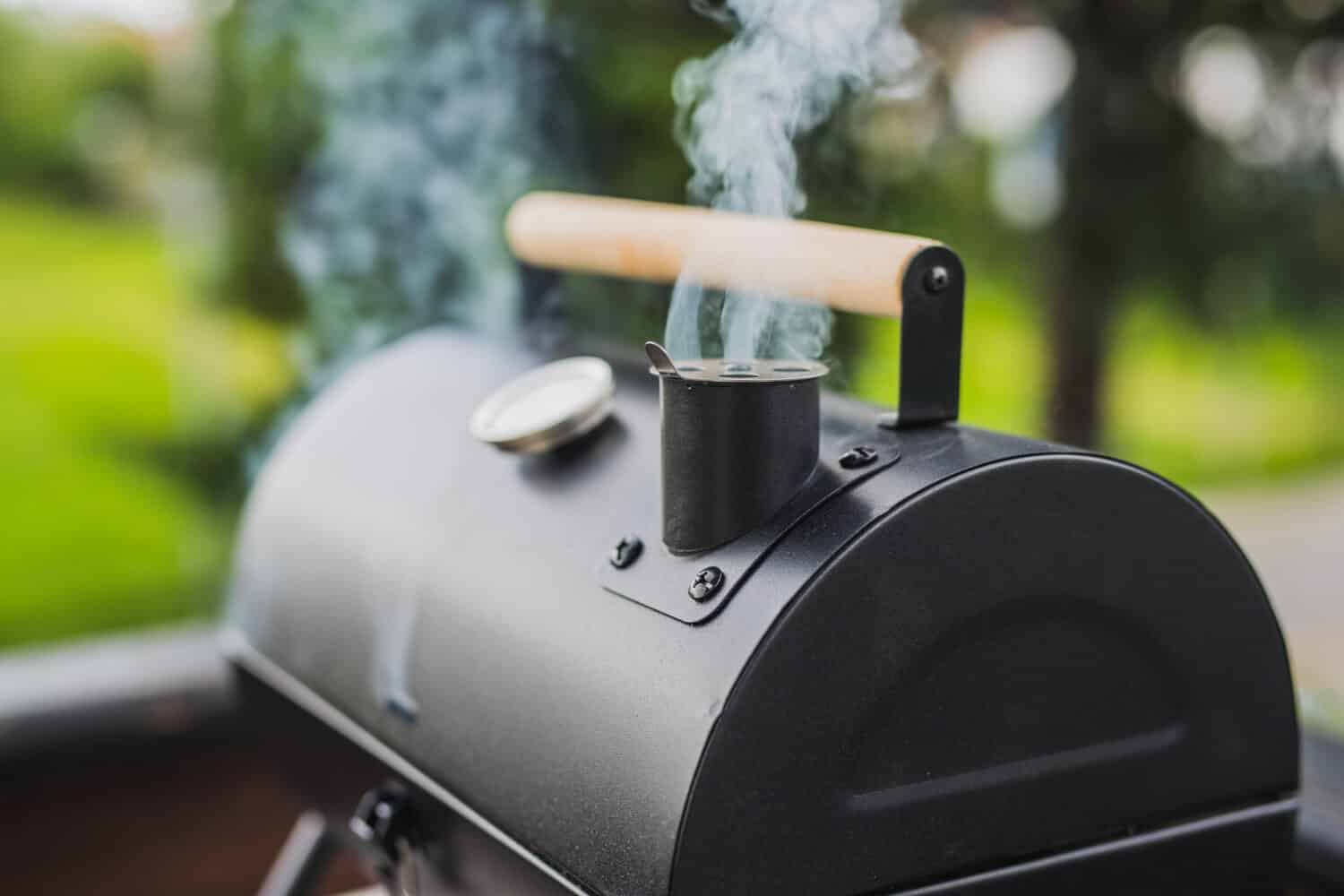 Smoke coming out of a smokestack of a small black smoker grill or barbecue on green background.