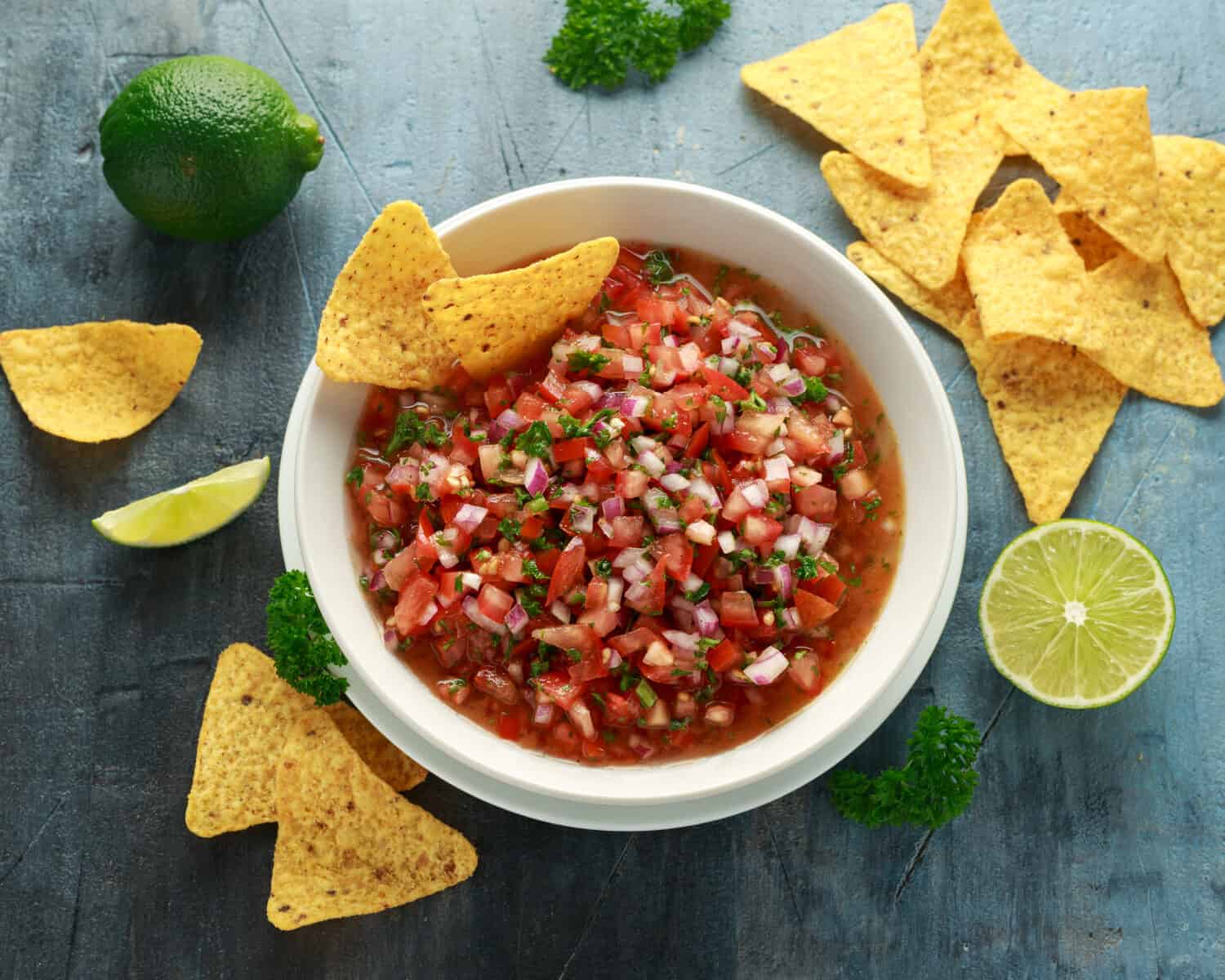 Mexican Tomato Salsa in white bowl with lime, red onion, jalapeno pepper, parsley and tortilla chips