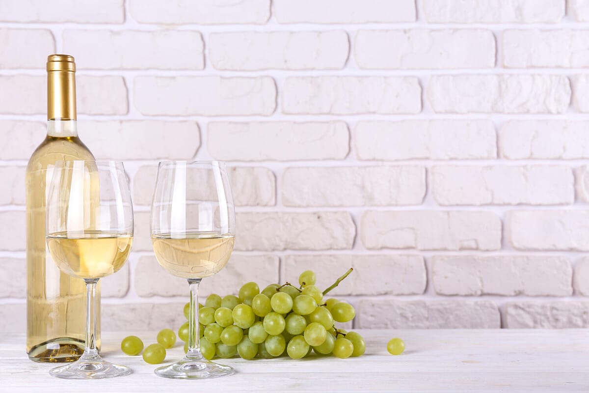 Vintage bottle of white wine without label, two glasses and bunch of grapes on wooden table, lofty white brick wall background. Expensive bottle of chardonnay concept. concept. Copy space.