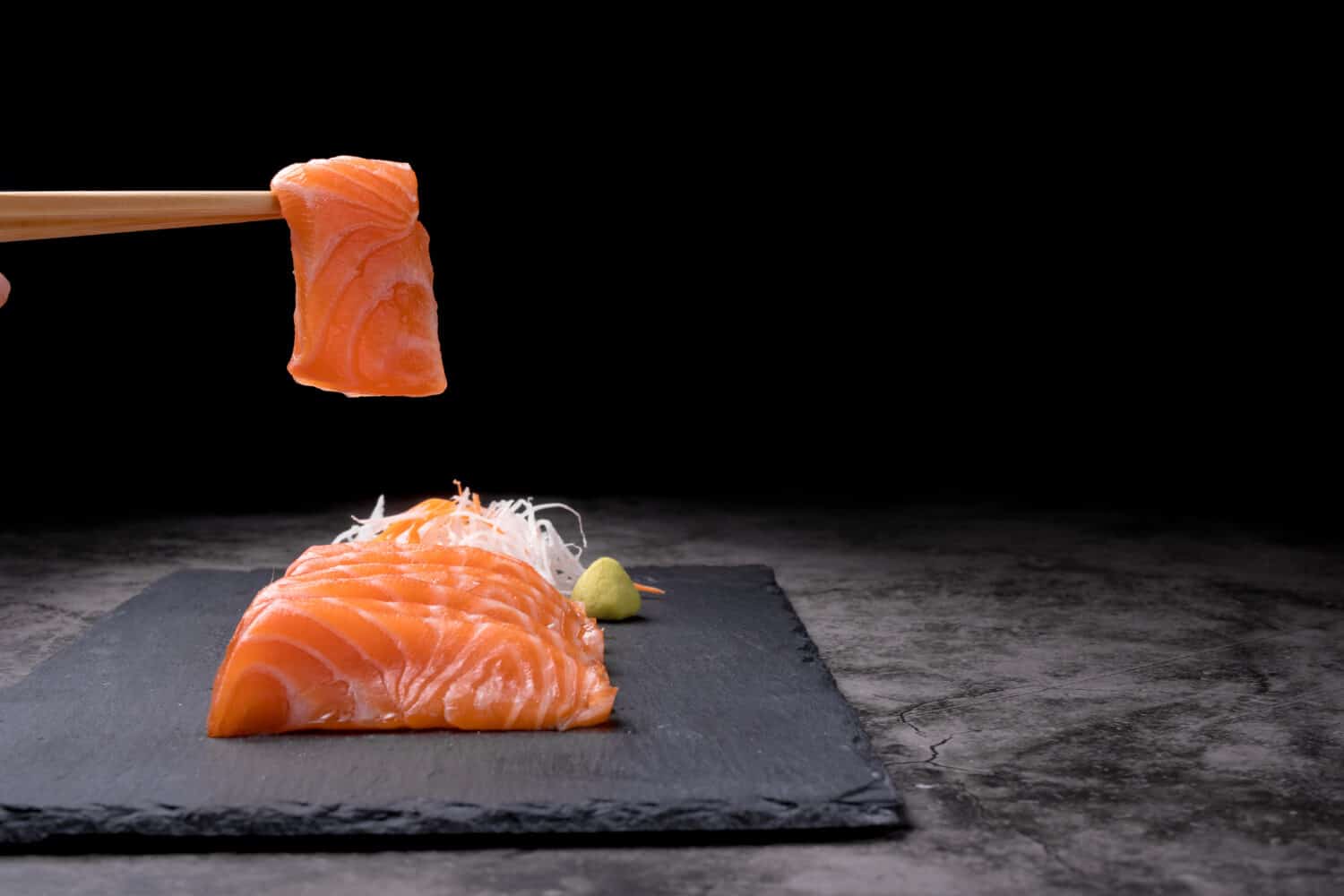 The hands were holding the chopsticks to hold the salmon sashimi, which was arranged on a black stone plate on a old table, with copy space.