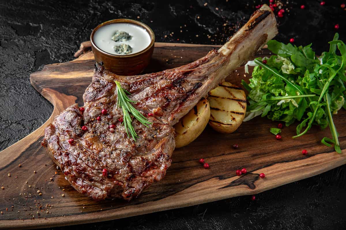 Juicy delicious ribeye or tomahawk steak on the bone with baked potatoes, spices and herbs. Hearty meat dish