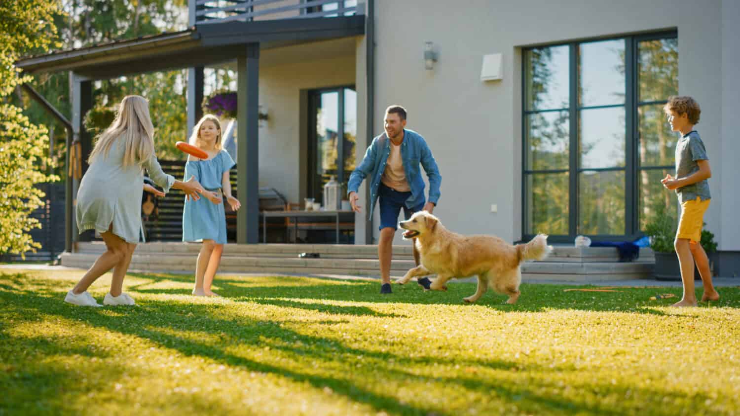 Smiling Beautiful Family of Four Play Fetch flying disc with Happy Golden Retriever Dog on the Backyard Lawn. Idyllic Family Has Fun with Loyal Pedigree Dog Outdoors in Summer House Backyard