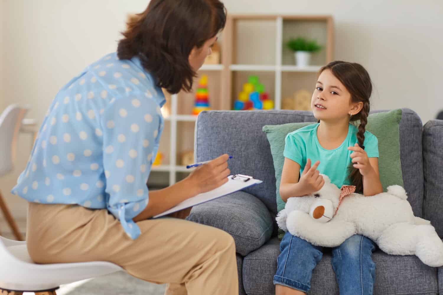 Supportive psychologist with clipboard listening to little child during therapy session. Preschool girl feeling at ease in therapist's office sharing her thoughts and concerns