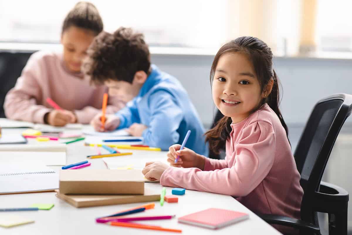 Academic Concept. Smiling junior asian school girl sitting at desk in classroom, writing in notebook, posing and looking at camera. Group of diverse classmates studying in the background