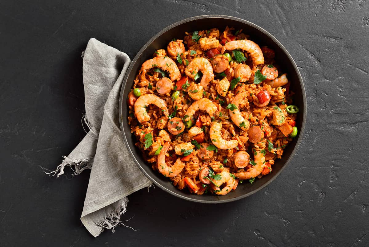 Creole style jambalaya with prawn, chicken, smoked sausages and vegetables in frying pan over black stone background. Top view, flat lay, close up