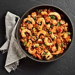 Creole style jambalaya with prawn, chicken, smoked sausages and vegetables in frying pan over black stone background. Top view, flat lay, close up