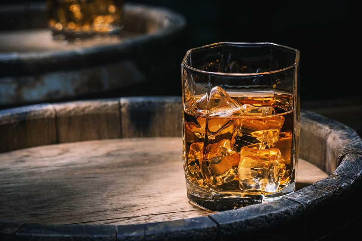 Bottle and glass of whiskey with ice on a wooden background. Glass of Scotch whiskey and ice sits on top of a rustic whiskey barrel. Whiskey with ice.