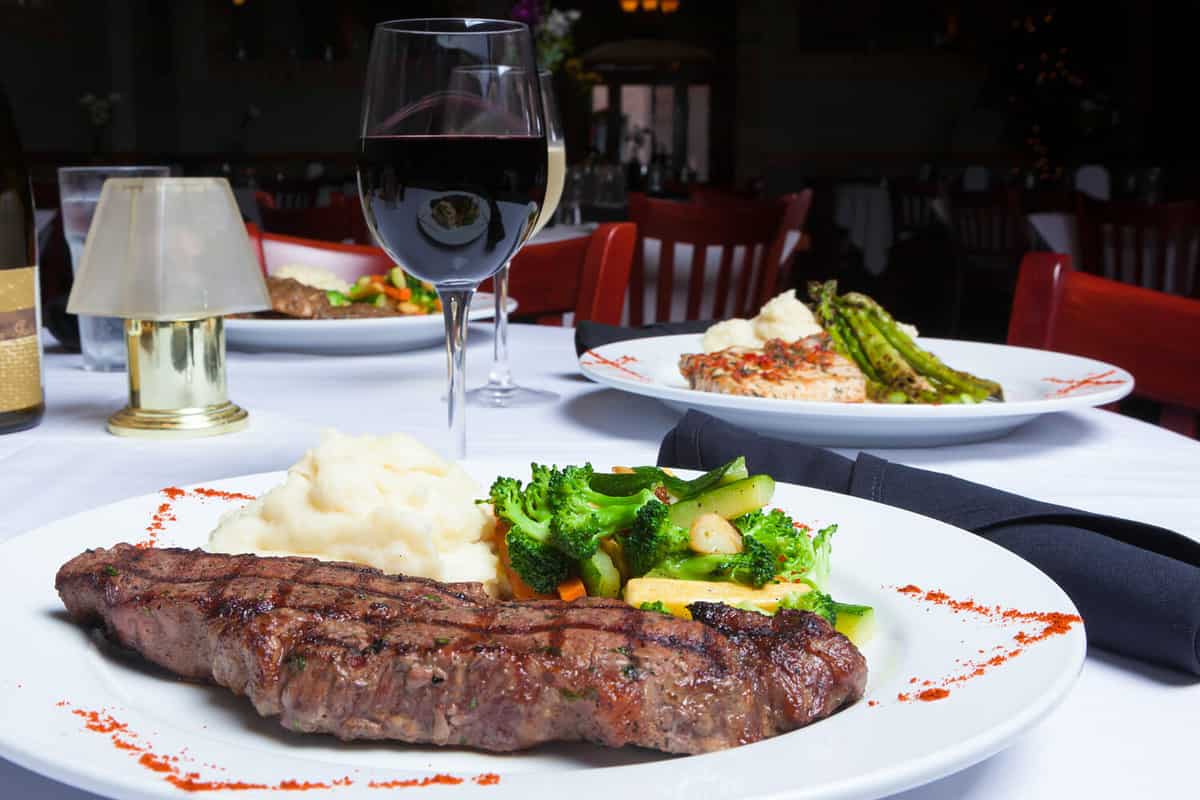 A white plate holds a delicious grilled New York Strip Steak with a side of mashed potatoes and fresh mixed vegetables. A glass of red wine completes the meal.