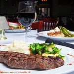 A white plate holds a delicious grilled New York Strip Steak with a side of mashed potatoes and fresh mixed vegetables. A glass of red wine completes the meal.