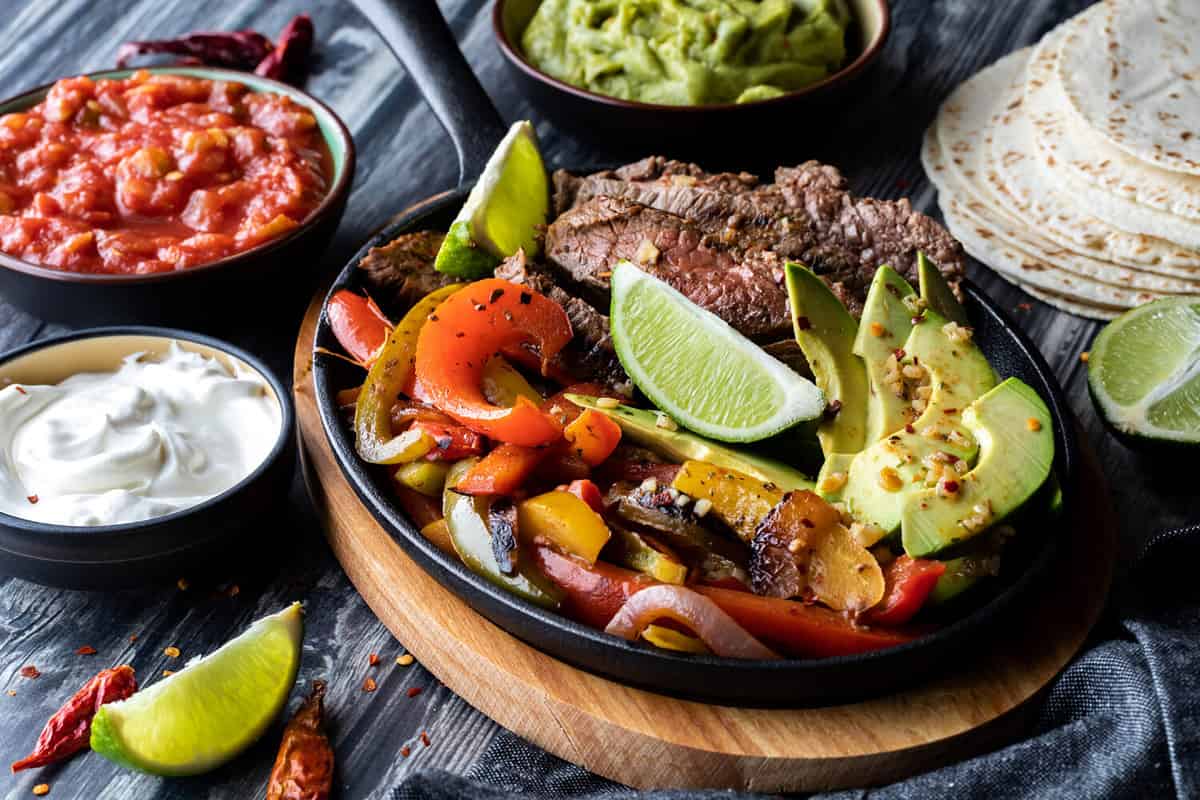 Close up of a cast iron skillet filled with fajita ingredients and surrounded by more toppings and tortilla wraps.