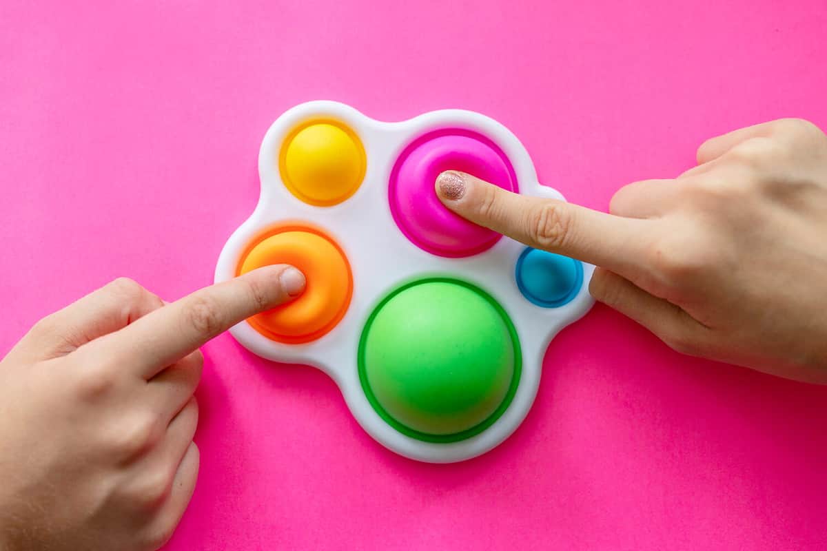 colorful trendy antistress sensory toy fidget push pop it and simple dimple in kid's hands, children share toys and playing together on pink background