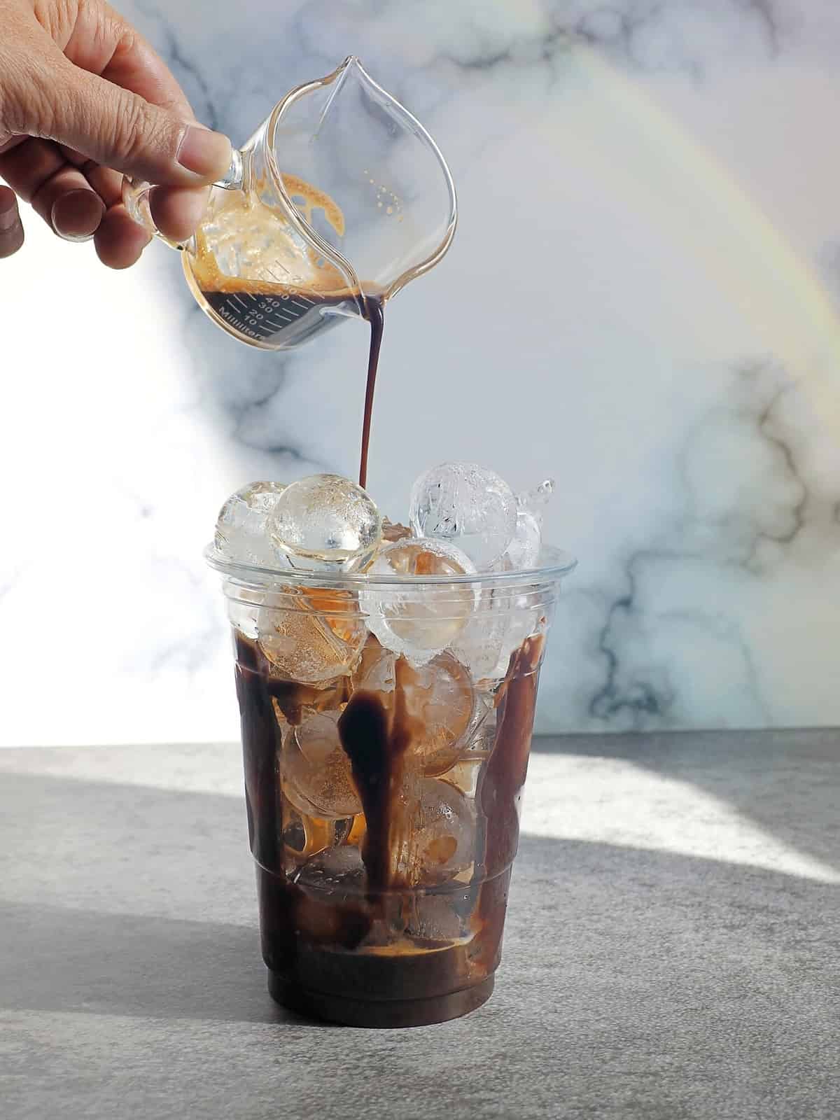 Hand pouring espresso over ice balls in disposable transparent plastic glass on rustic grey background and white marble wall with rainbow light reflection. Enjoy making summer drink theme.