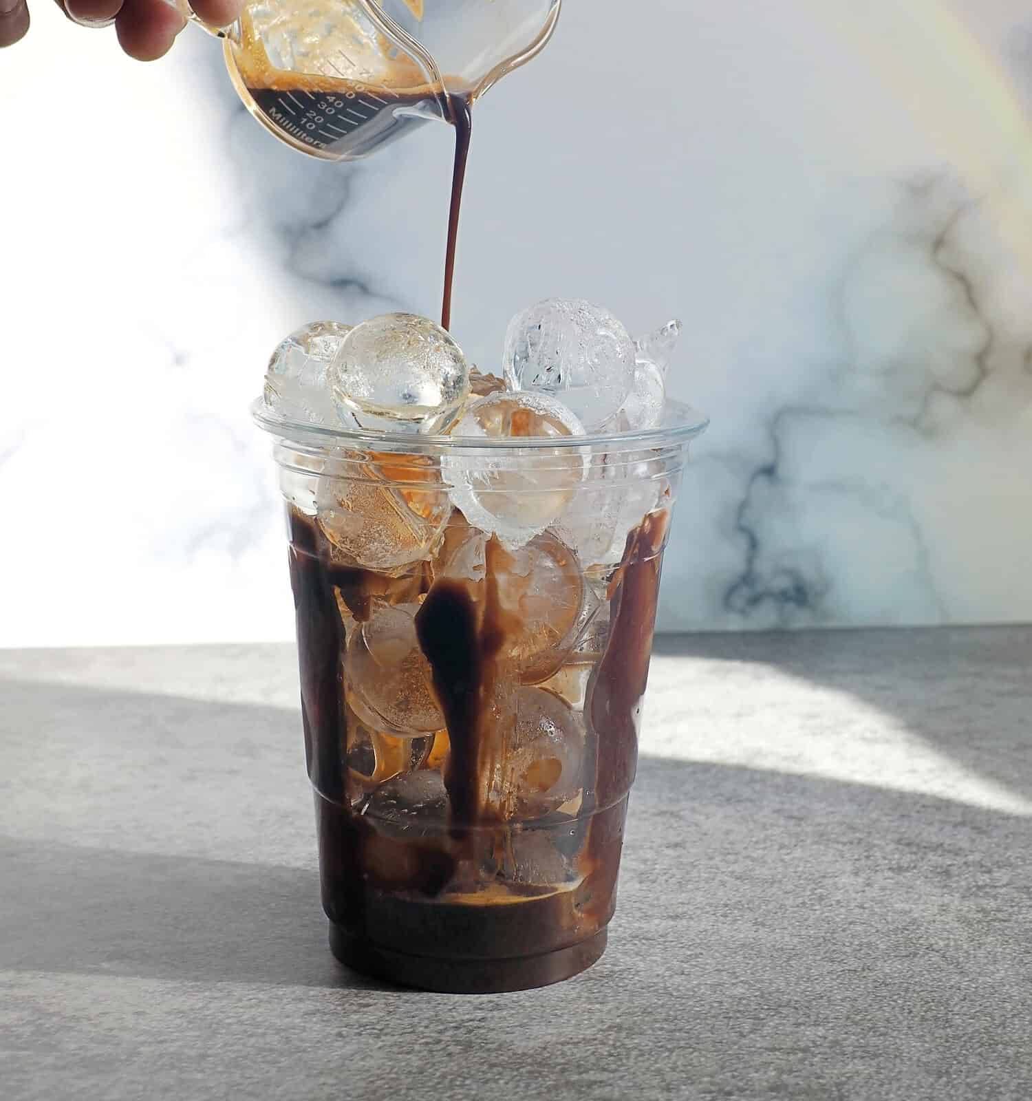 Hand pouring espresso over ice balls in disposable transparent plastic glass on rustic grey background and white marble wall with rainbow light reflection. Enjoy making summer drink theme.