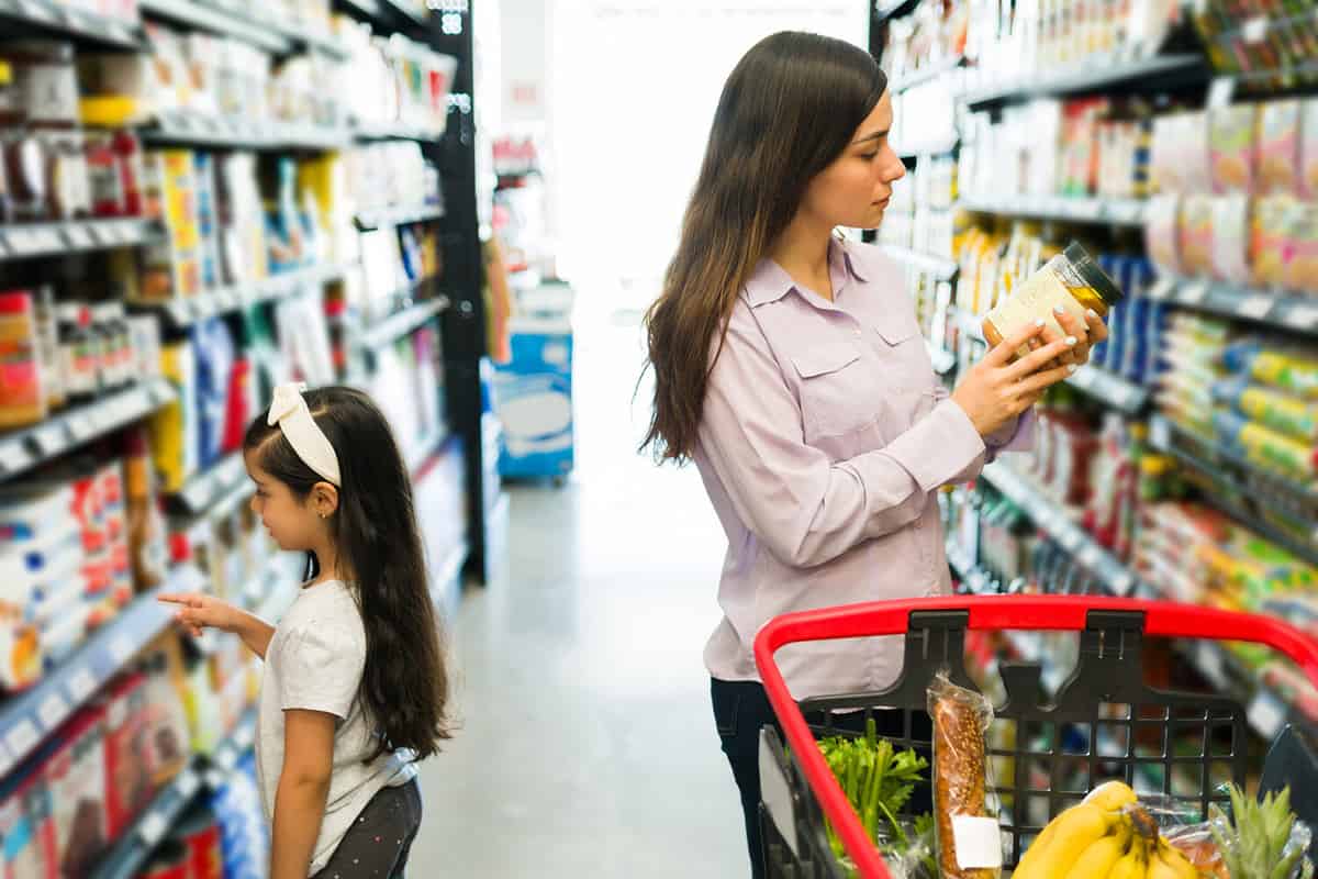 Side view of an hispanic mother buying products at the grocery store with her daughter and pushing a shopping cart on the supermarket aisle