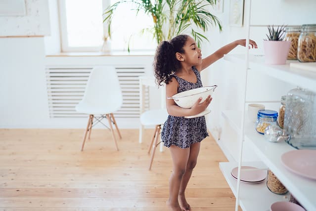 Side view of black girl with curly pigtails and dirty face in cute blue dress holding strainer and reaching hand on kitchen dish shelf in white room
