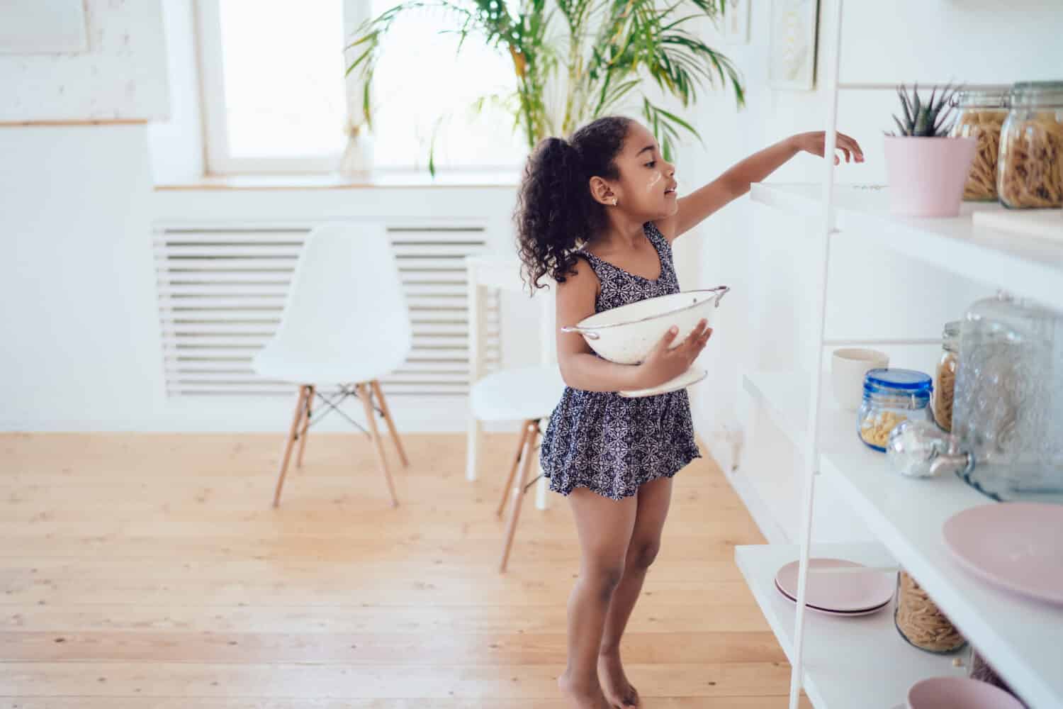 Side view of black girl with curly pigtails and dirty face in cute blue dress holding strainer and reaching hand on kitchen dish shelf in white room