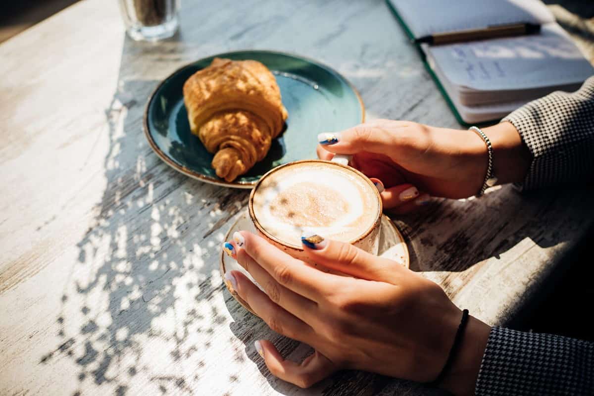 Cappuccino and croissant on the table in the cafe. The morning sunlight falls on the table, beautiful shadows appear. Delicious breakfast.Women's hands hold a cup of coffee.