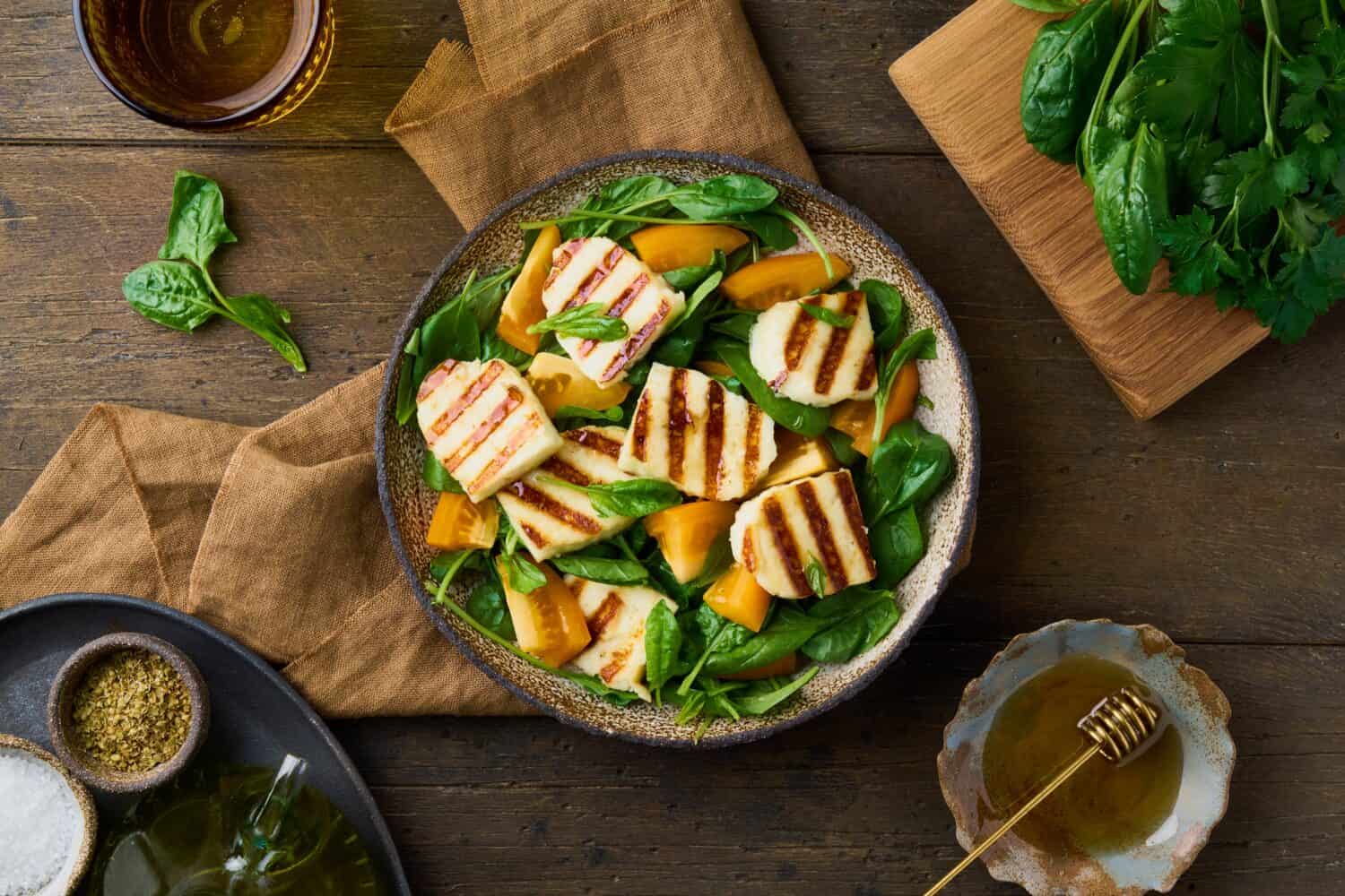 Tasty vegetarian healthy salad with orange tomatoes, fresh spinach, grilled halloumi cheese, honey and olive oil sauce with spices. Horizontal, top view, brown wood background