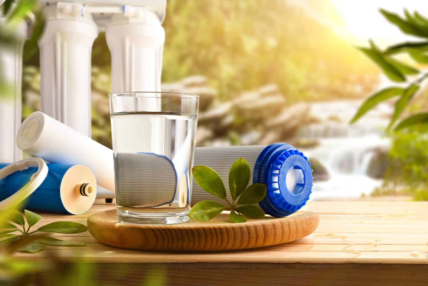 Glass of water purified by osmosis with environmentally friendly biodegradable filters on wooden slat table with nature background trees and waterfall. Front view. Horizontal composition.