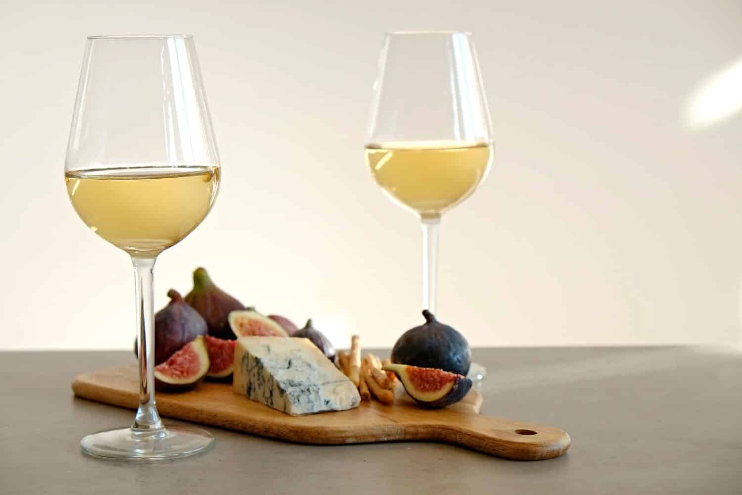 Two wineglasses of vintage chardonnay with delicious appetizers. Couple of glasses of white wine, italian breadsticks, figs and grapes. Interior background. Close up, copy space.