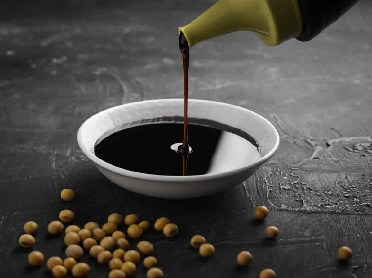 Pouring sweet soy sauce into white ceramic bowl from bottle on black background. soybean seeds in frame