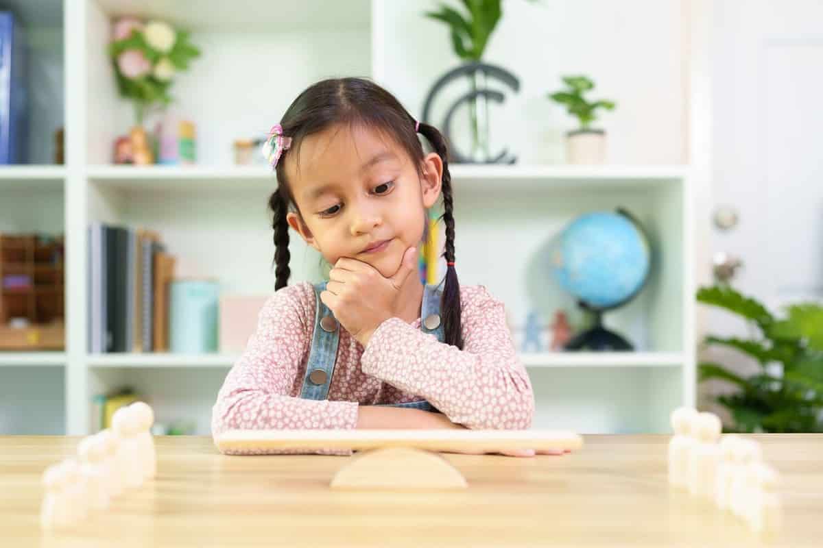 Cute asian 6 years old girl is thinking how to balance the wooden people model on the scale, concept of learn through play, montessori, homeschool, creativity, stem education and child development.