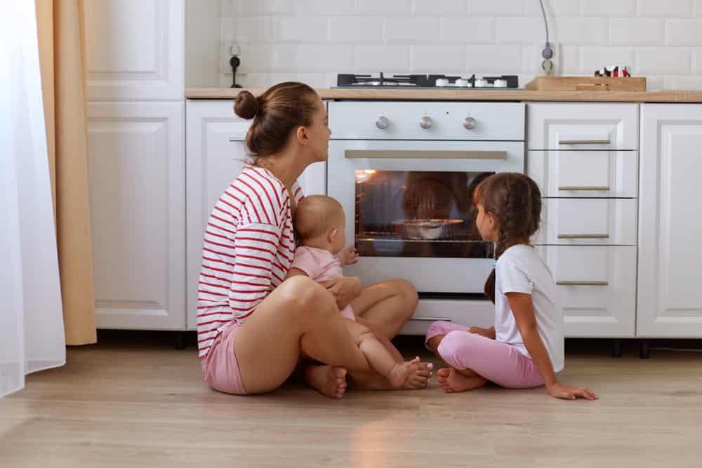 Horizontal shot of happy woman with bun hairstyle wearing striped shirt sitting on floor in kitchen with her kids, mother and her kids waiting for tasty pie, baking in oven.