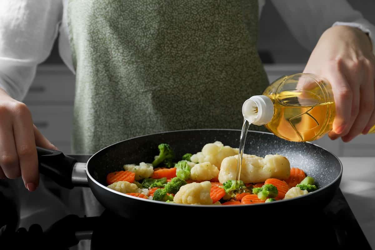 Woman pouring cooking oil from bottle into frying pan with vegetables on stove, closeup