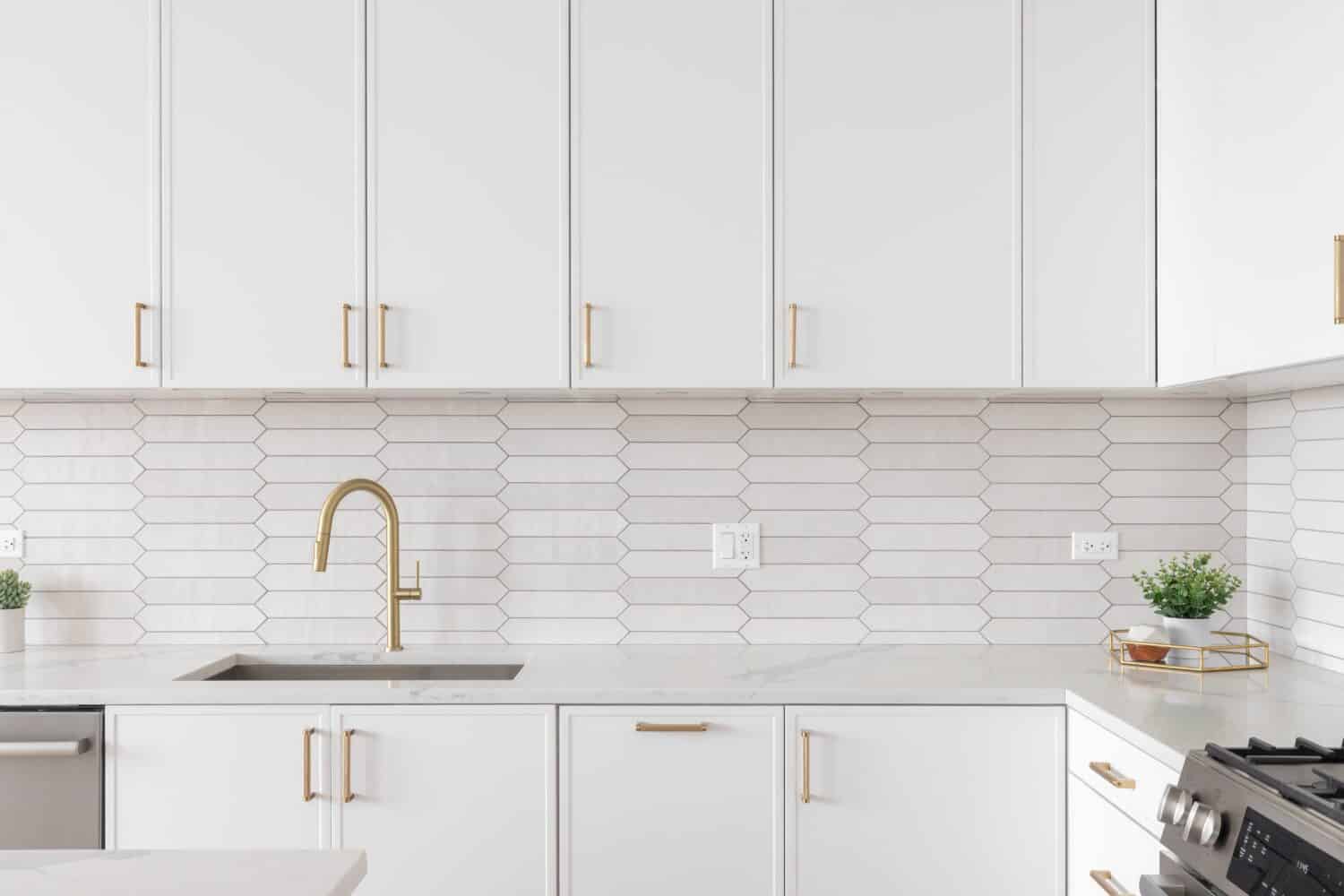 A beautiful kitchen faucet detail with white cabinets, a gold faucet, white marble countertops, and a brown picket ceramic tile backsplash.