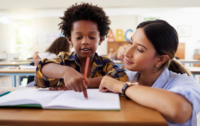 Teacher, learning and helping black kid in classroom for knowledge, studying or assessment. Question, development and boy or student with woman for education pointing in notebook in kindergarten.