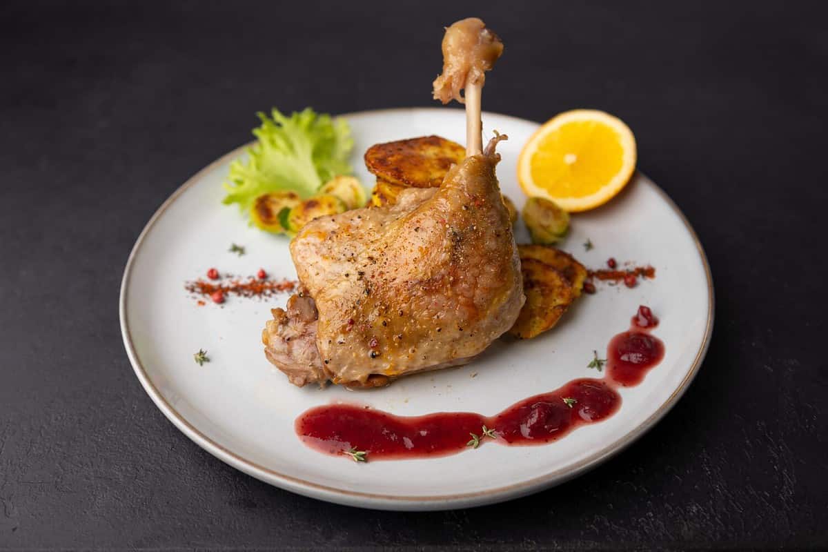 Duck leg confit with Brussels sprouts, baked potatoes, thyme, orange and lingonberry sauce. Traditional French cuisine. Selective focus, close-up, black background.