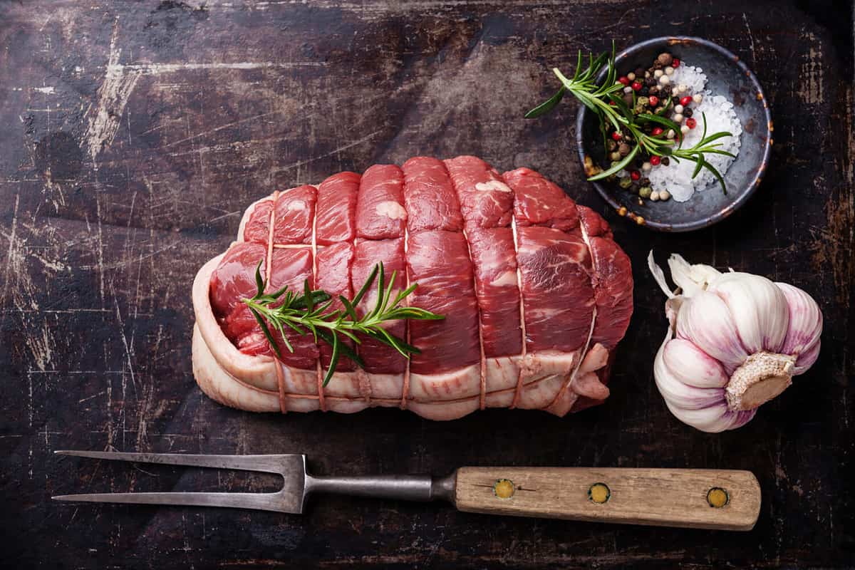 Raw roast beef Rump, seasonings and meat fork on dark metal background