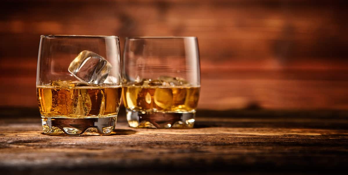 Two glasses of whiskey with ice cubes served on wooden planks. Vintage countertop with highlight and a glass of hard liquor