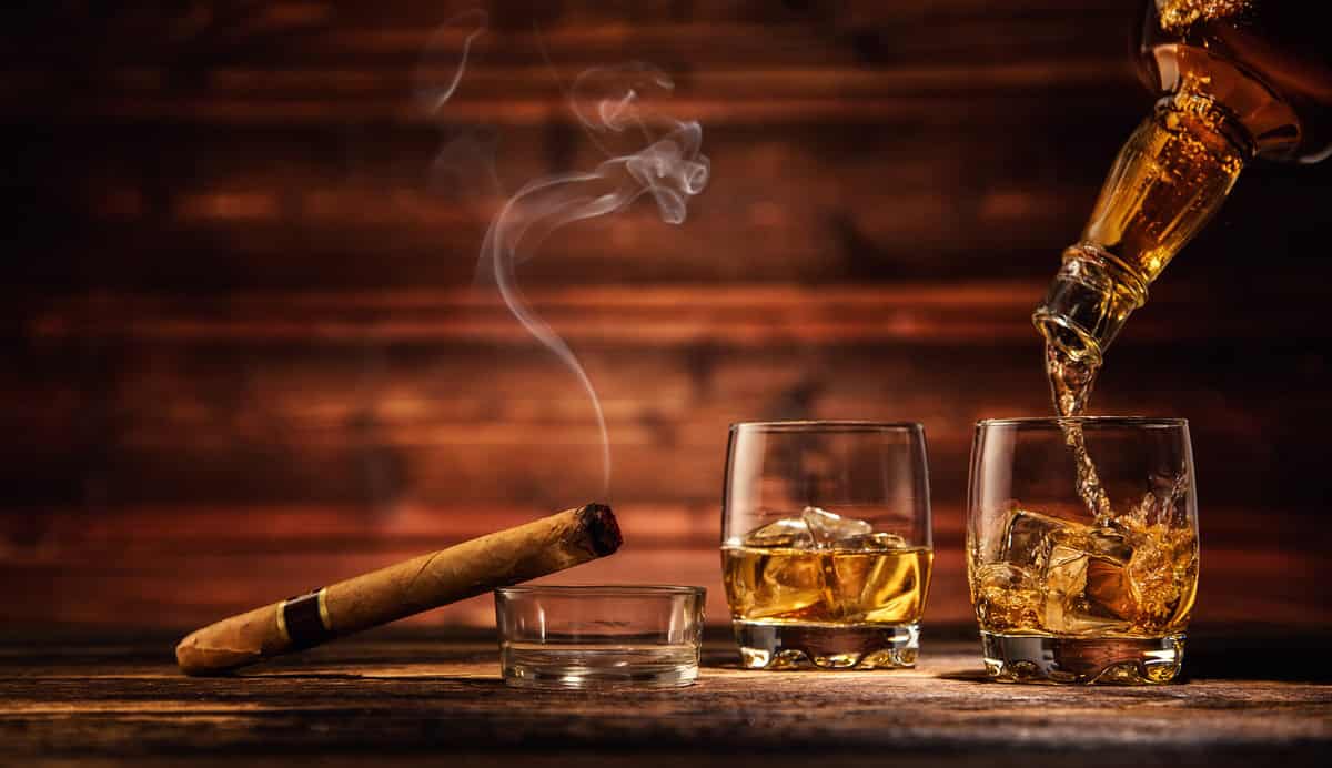 Pouring whiskey from bottle to two glasses with ice cubes, served on wooden planks. Vintage countertop with highlight and a glass of hard liquor