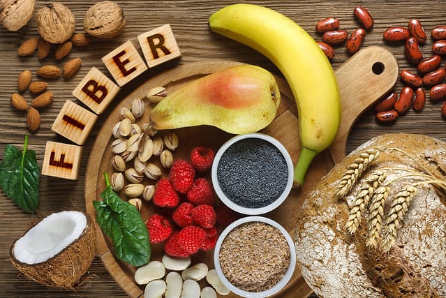 Foods rich in fiber as rye bread, wheat bran, white beans, red beans, spinach, almonds, poppy seed, pears, bananas, coconut, raspberries, pistachios, walnuts. Wooden table as background