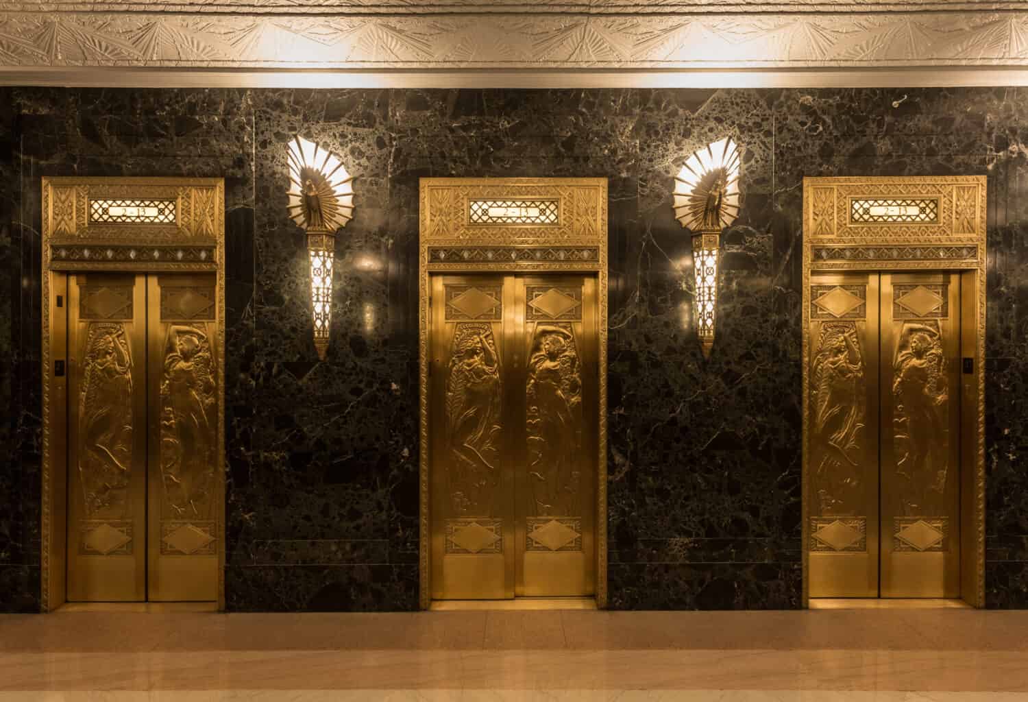 Dramatic golden elevators in fancy urban skyscraper building with relief sculptures and marble wall with lights and sconces 