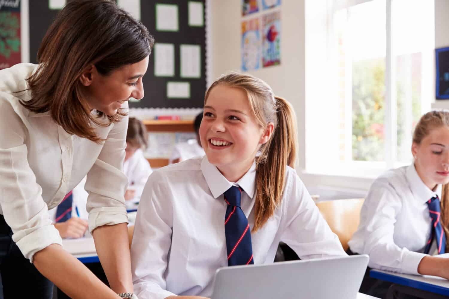 Female Teacher Helping Pupil Using Computer In Classroom