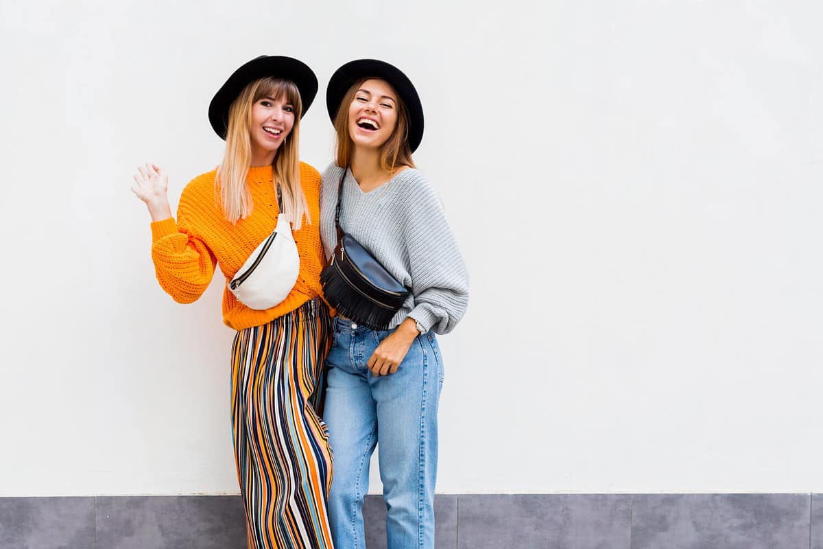 Two exited hipster women posing over white wall. Fall season. Wearing stylish orange knitted sweater, black hat , bum bag. Friends spending great time together . Copy space .