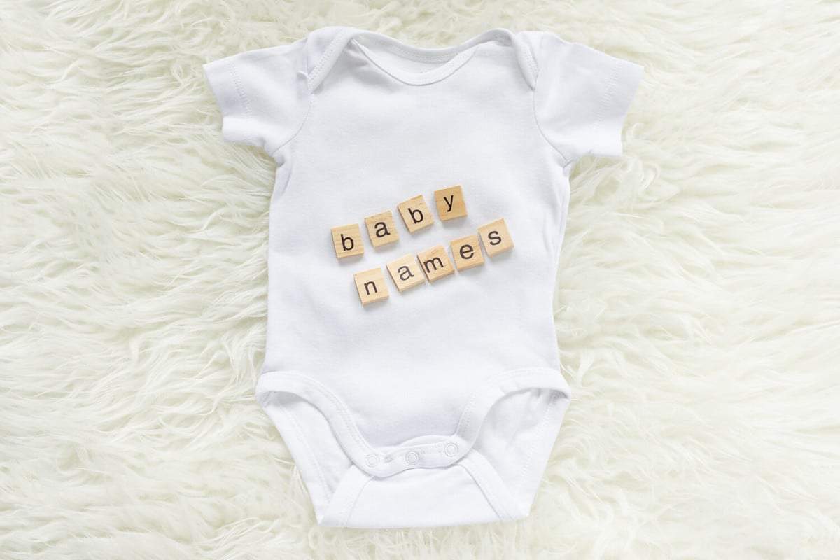 White baby bodysuit with wooden tiles saying baby names on a fluffy background