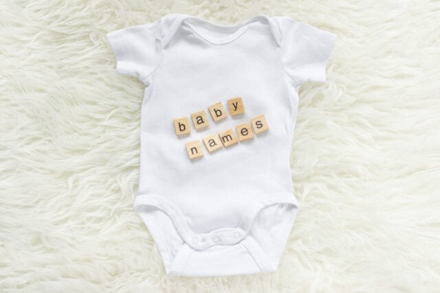 White baby bodysuit with wooden tiles saying baby names on a fluffy background