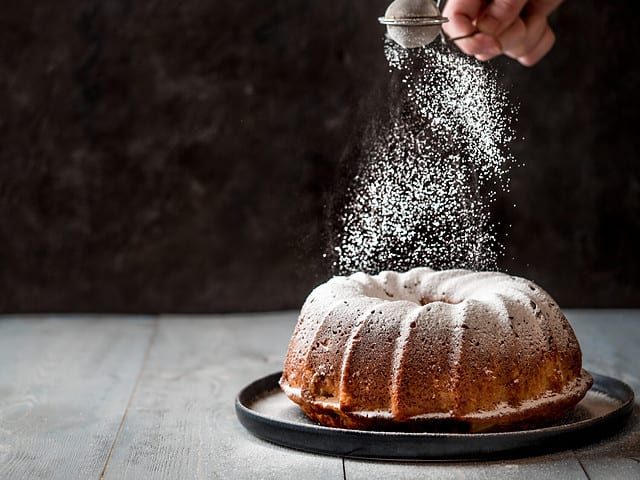 Woman's hand sprinkling icing sugar over fresh bundt cake. Powder sugar falls on fresh perfect bundt cake. Copy space for text. Ideas and recipes for breakfast or dessert. Black background