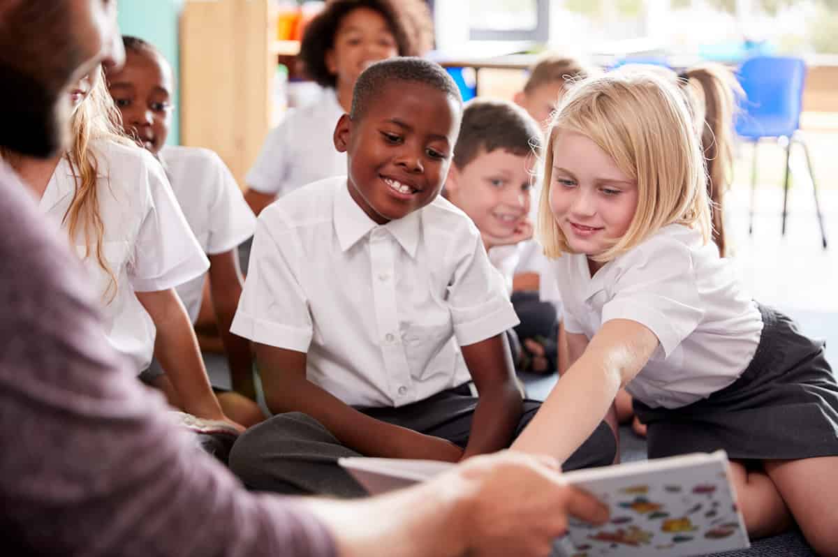 Male Teacher Reading Story To Group Of Elementary Pupils Wearing Uniform In School Classroom