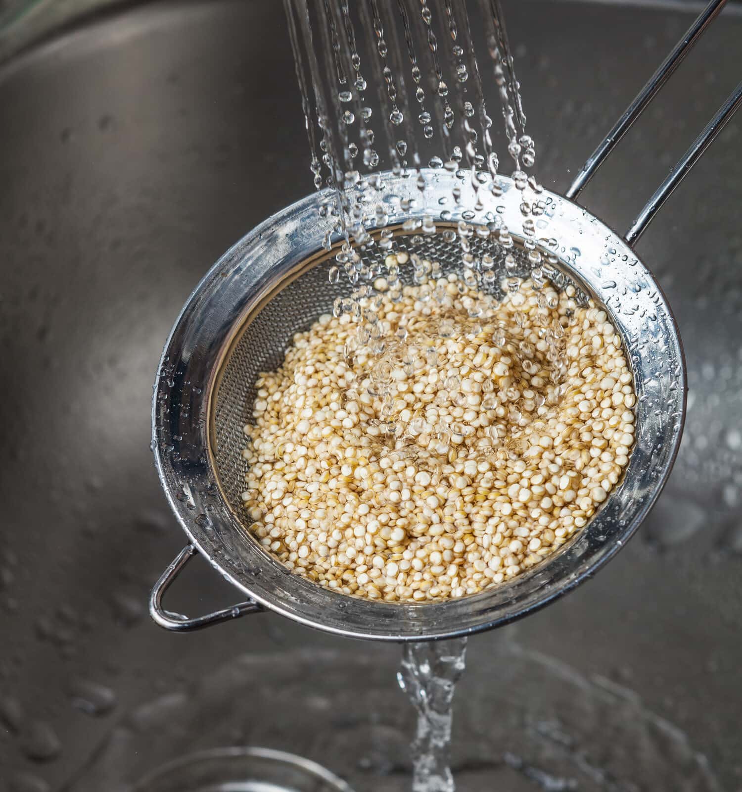 Small strainer with raw quinoa seeds and rinsing it in cold water in the kitchen sink