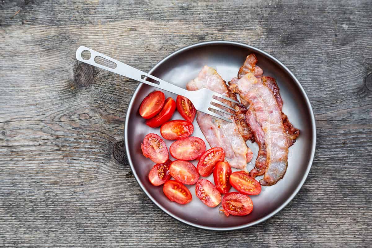 Breakfast, bacon,tomatoes and fork on a titanium plate. Tourism in the summer forest. Camping concept.