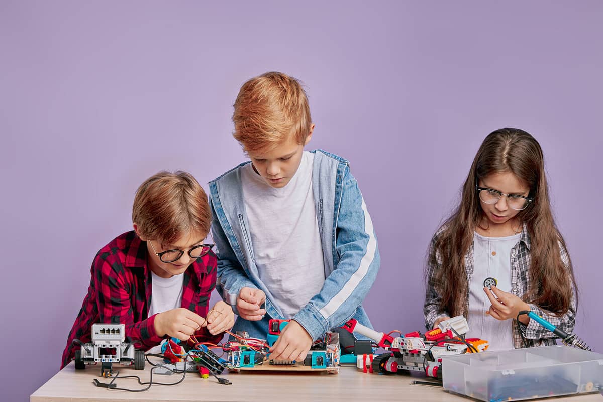 portrait of little 6-8 years old children stand together near table and assemble robotics isolated over purple studio, background. wearing casual wear