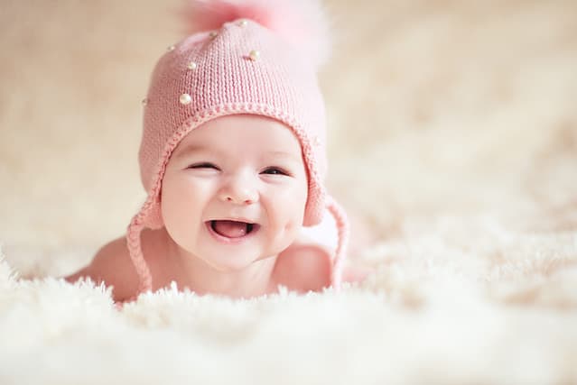 Laughing baby under 1 year old wearing knitted pink hat lying in bed closeup. Looking at camera. Happiness. Childhood.