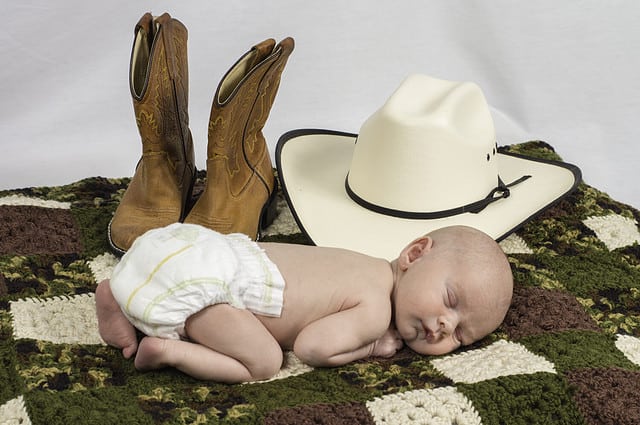 An infant baby laying down surrounded by a cowboy hat and boots.