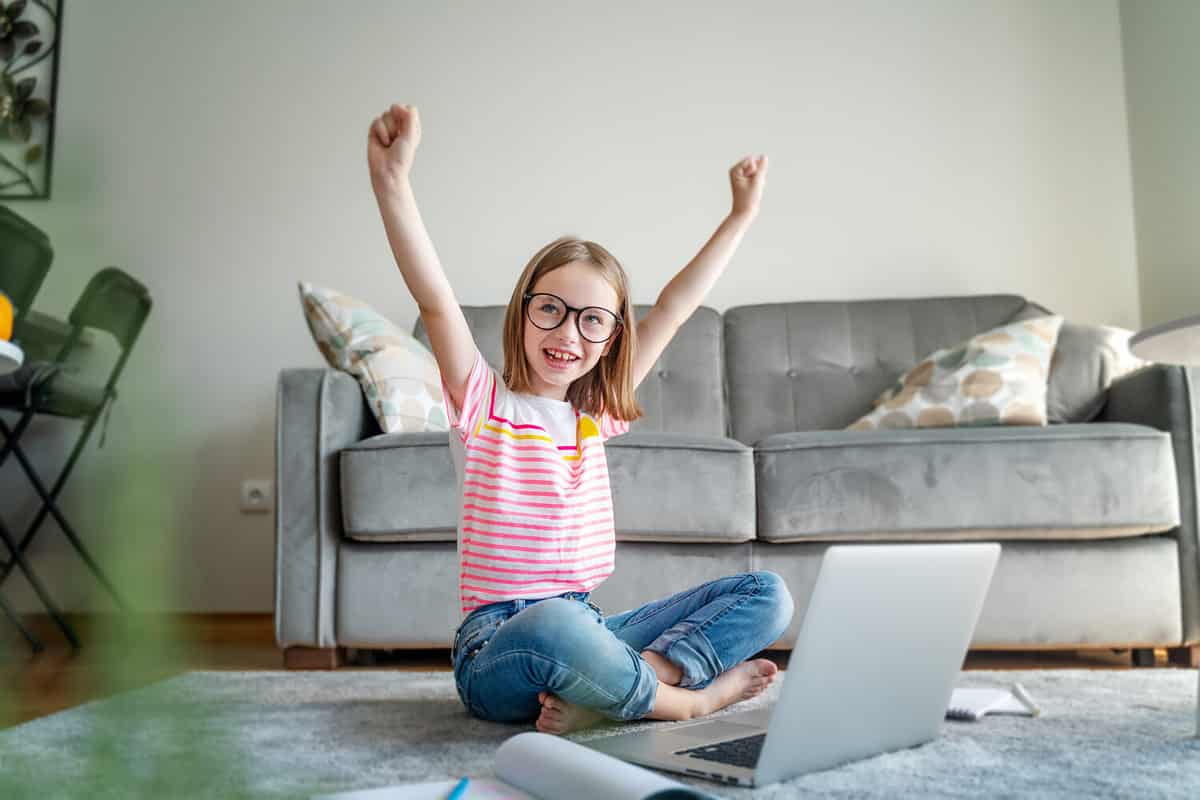 Happy cute little girl 8 years old in a striped t-shirt and jeans with glasses sits at home on a carpet in front of a laptop, remote education technologies and homework