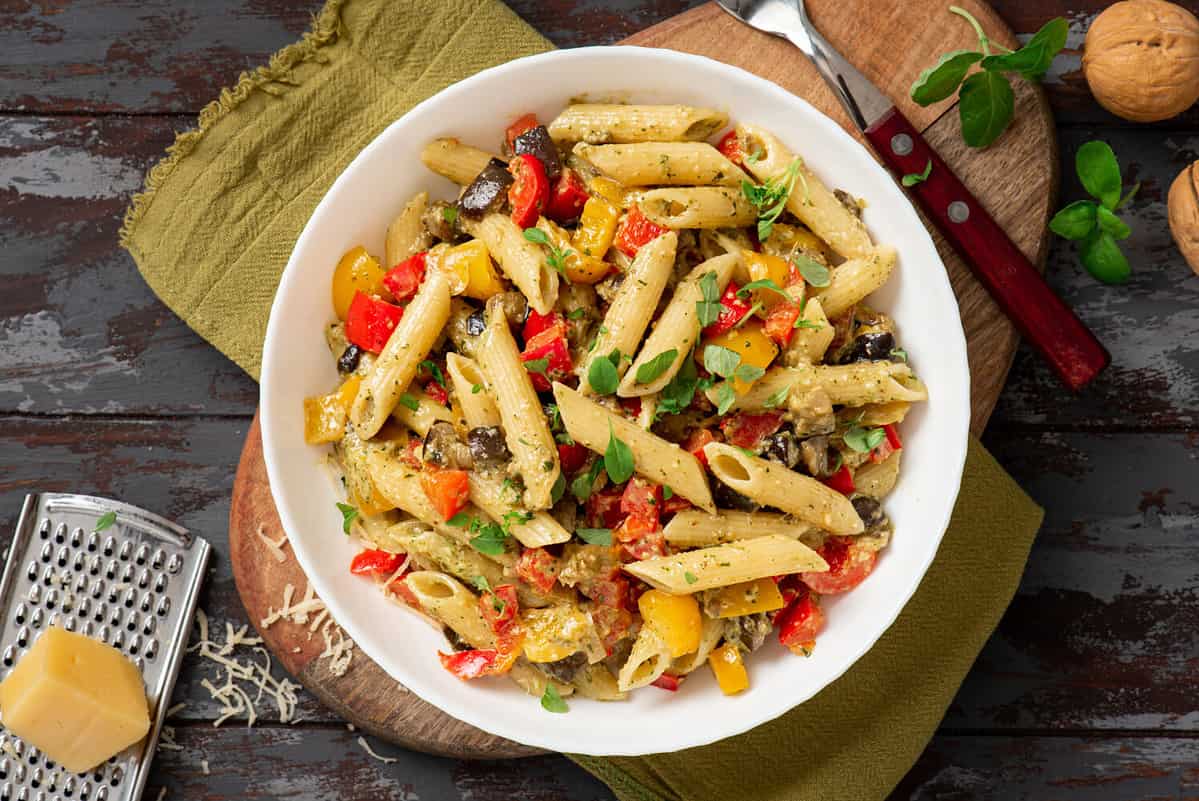 Pasta salad with baked vegetables. Penne pasta with baked peppers, eggplant, pesto and cheese in a white plate on a dark wooden table top view. Italian food. Rustic style.